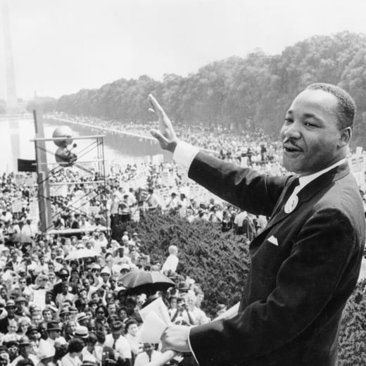 A gray scale photo of Martin Luther King Jr. addressing a large crowd from stage.